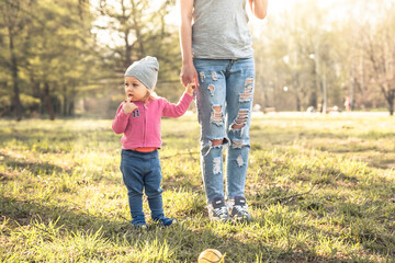 Fototapeta premium Child with mother standing together with holding hands in summer park on grass. Main subject is child. Unrecognizable mother on photo. Concept for togetherness and care.