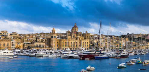 Fototapeta na wymiar Senglea, Malta - Panoramic vew of yachts and sailing boats mooring at Senglea marina in Grand Canal of Malta on a bright sunny summer day woth blue sky and clouds