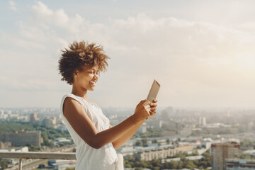 Laughing beautiful afro american teenage girl is making panorama image on her digital tablet while standing on observation point of high building with cityscape in blackground on warm sunny day - Powered by Adobe