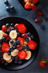 Useful breakfast of fruits and berries, top view. Sliced bananas, strawberries, grapes and blueberries in a black plate