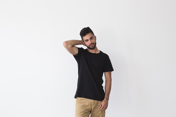 Smiling brutal spanish man with three day beard smirks and smiles into camera  wears blank black t-shirt, chino pants for summer season and friendship bracelet isolated on white mockup