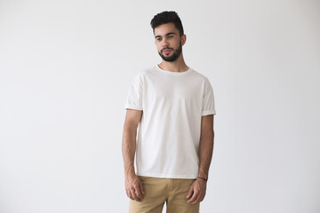 Fototapeta premium Front shot of hispanic or caucassian attractive hipster millennial male model softly smiling and wearing simple white blank t-shirt on isolated mockup background
