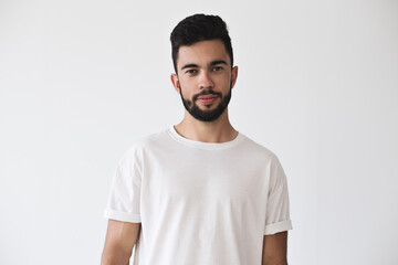 Portrait of attractive handsme young millennial with beard who looks into camera on isolated simple background and wears white t-shirt from premium organic materials