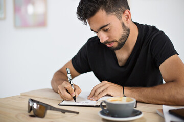 Attractive bearded student works on homework or art project in stylish hipster cafe full of light, sips on artisanal speciality coffee cappuccino