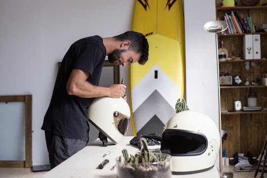 Handsome smiling artist decorates full face motorcycle helmet with thick oil brush and ink marker, inside designer interior industrial loft or art studio in hip district