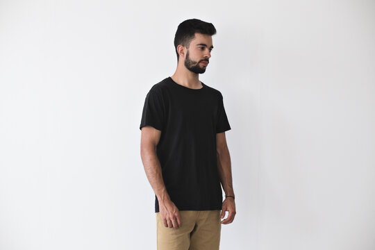 Bearded hipster youngster in studio setting poses in blank black t-shirt and chinos trousers, friendship bracelet softly smiles into the camera, calm and tranquil, on white mockup