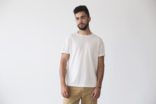 Front shot of hispanic or caucassian attractive hipster millennial male model softly smiling and wearing simple white blank t-shirt on isolated mockup background