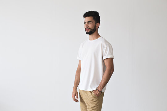 Attractive bearded masculine male poses with hand in his pocket on isolated mockup wall in chinos pants and simple yet stunning white t-shirt made from organic cotton