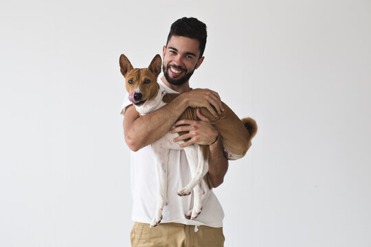 Happy laughing attractive male holds and hugs his best friend, little basenji brown puppy dog who is licking his face after tasty meal