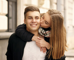 Young happy joyous couple cuddling at city street. Beautiful pretty girl sitting at man's back and embracing him.