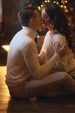 Young Couple Staying At Home Together On Holiday Evening And Sitting On Floor In Front Of Christmas Tree And Hugging. Cute Romantic Lovers In White Pullovers