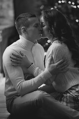 Young couple staying at home together on holiday evening and sitting on floor in front of christmas tree and hugging. Cute romantic lovers in white pullovers