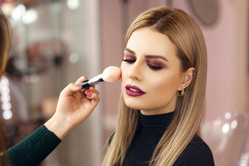 Process of making makeup. Mak-up artist working with brush on model face. Portrait of young blonde woman in beauty saloon interior. Applying tone to skin.