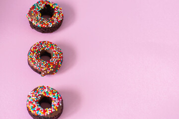 Three baked chocolate doughnuts with chocolate glaze with confetti over pink background. Overhead view.