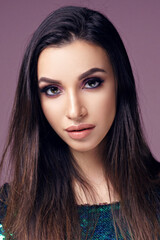 Fototapeta premium Closeup beauty studio fashion portrait of young pretty girl with long brunette dark hair and nakeup posing in shiny green dress with sequins posing against purple background and looking in camera.