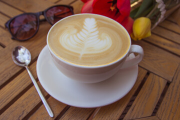 Cup of hot latte art coffee, glasses and tulips on wooden table
