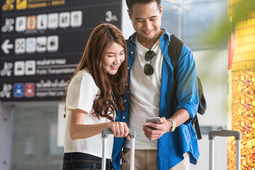 Asian couple traveler using the smart mobile phone for check-in at the flight information screen in moddern an airport, travel and transportation with technology concept.