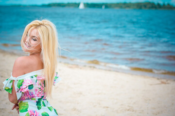 Fototapeta na wymiar Beautiful blonde hair woman in romantic flowers dress near blue water and sand. Cute happy girl have fun on a nature