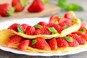 Strawberry omelette. Fried omelette filled with fresh strawberries and garnished with mint on a plate. Fresh strawberries on old wooden table. Sweet breakfast omelette recipe. Closeup