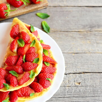 Summer berry omelette. Colorful easy omelette stuffed with fresh strawberries and garnished with mint on a plate and wooden background with empty copy space for text. Breakfast or lunch omelette idea