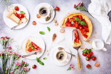 Overhead shot of cups of coffee, delicious homemade strawberry cheesecake and flowers on light gray background. Top view, flat lay.