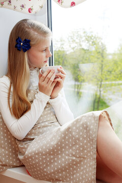 Beautiful child drinking tea and looking through window while sitting at windowsill at home