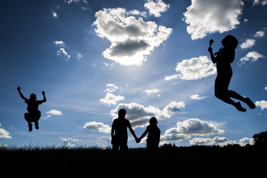 The silhouettes of  a women flying, a boy jumping and two young people holding hands  on the top one hill and the clear sky with some clouds