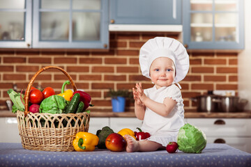 Smiling baby girl dressed in white chef toque and apron sits near wicker basket with fresh raw vegetables and applauds. Little scullion cooking in the kitchen. One year old child nutrition