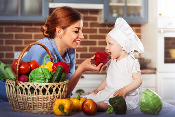 Smiling mother feeding charming one year baby girl cook. Little scullion dressed in white chef hat among different vegetables. Motherhood and childhood concept. One year old child nutrition.