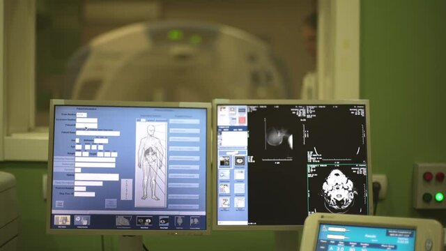 Crane motion, tilt up from monitor screen with x-ray images of human body in focus, patient behind protective glass lying on CT or MRI scanner in the blurred background, shallow depth of field