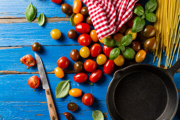 Tasty fresh appetizing variety of tomatoes, basil, spaghetti, kitchen towel and pan on vibrant blue wooden background. Top View. Cooking of italian pasta.
