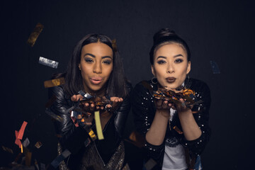 Beautiful stylish young women blowing shiny confetti isolated on black