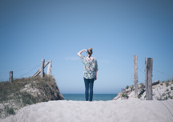 back view of blonde caucasian female standing on white sand of Baltic Sea beach shielding her eyes from the sun with one hand while looking at the water