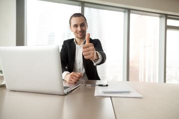 Fototapeta na wymiar Confident businessman showing thumbs up gesture at desk in office. Entrepreneur satisfied with work results. CEO supports good decision, recommends proven solution. Company leader like successful idea