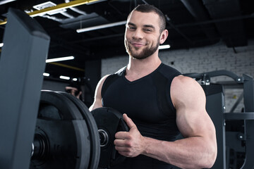 Fototapeta na wymiar Smiling muscular man putting weight on dumbbell and looking at camera in gym