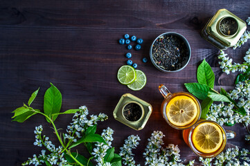 Theme of tea, background with space for text, dark. Black tea in transparent mugs with lemon, ginger, lemons, limes, mint, cherry blossoms. Food industry, tea packing, restaurant business
