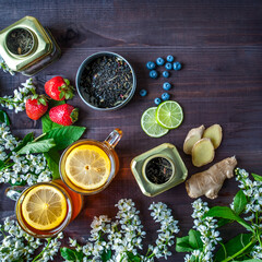 Theme of tea, background with space for text, dark. Black tea in transparent mugs with lemon, ginger, lemons, limes, mint, cherry blossoms. Food industry, tea packing, restaurant business
