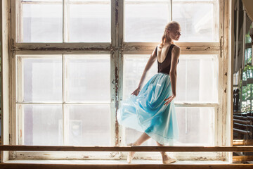 A ballerina is posing in front of a large window, standing on the windowsill. Girl in a turquoise ballet skirt.