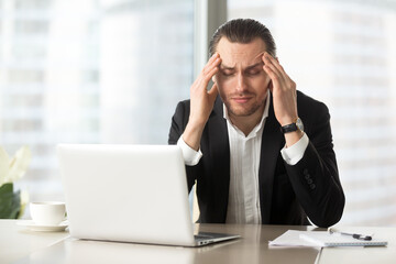 Frustrated man squeezes temples with hands at desk in front of laptop. Tired businessman suffers from headache at workplace. Entrepreneur feels chronic fatigue, work stress because of business problem