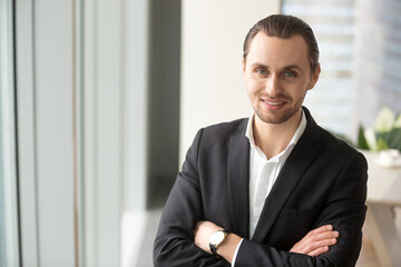 Portrait of young successful company leader standing with arms crossed and looking in camera with smile. Entrepreneur confident in his own abilities posing in office interior. Satisfied by success CEO