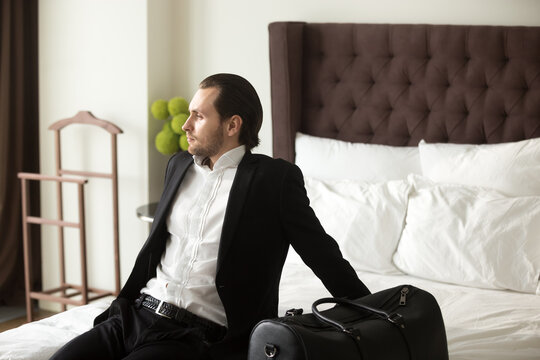 Man in business suit sitting on hotel room bed with luggage, relaxing, looking in window. Successful businessman ready to business trip abroad, dreaming about weekend leisure, foreign country vacation