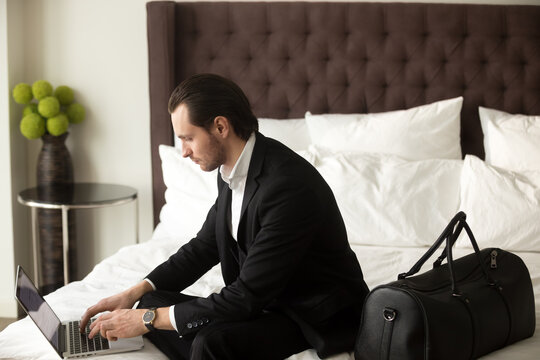 Serious man wearing business suit working on laptop while sitting on bed with luggage in luxury apartment. Businessman finishing presentation in hotel room before going on meeting with foreign partner