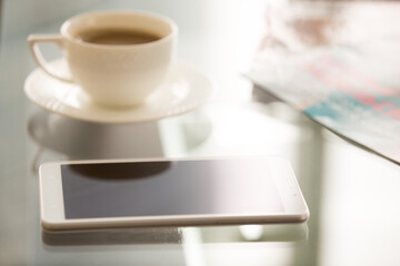 Fototapeta na wymiar Close up image of trendy smartphone on glass desk surface near white porcelain cup. Shiny cellphone lying on coffee table. Take a break with mobile phone in cafe concept. Fresh coffee before work