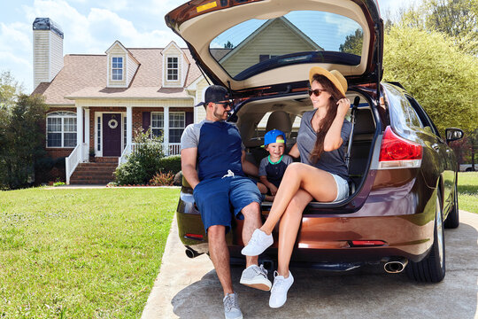 Family sits cars trunk near house. Mother, father, son.