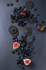 Variety of fresh berries blueberry, dewberry and figs with black charcoal crackers over dark metal background. Top view with space. Summer dark snacks concept