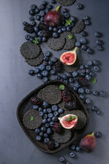 Variety of fresh berries blueberry, dewberry, red currant and figs with black charcoal crackers on wooden plate over dark metal background. Top view with space. Summer dark snacks concept