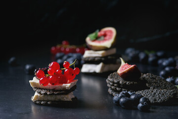 Black charcoal crackers with camembert brie cheese and berries blueberry, dewberry, red currant and sliced figs, served on vintage tray over dark metal background. Summer appetizer