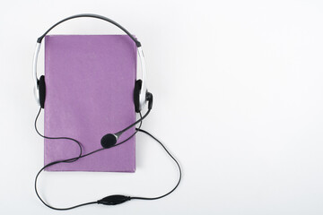 Audiobook on white background. Headphones put over purple hardback book, empty cover, copy space for ad text. Distance education, e-learning concept.