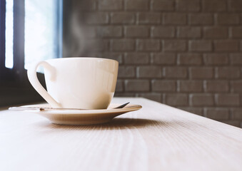 Coffee cup on wooden table against brick wall background with smoke and window lighting. Selective focus.
