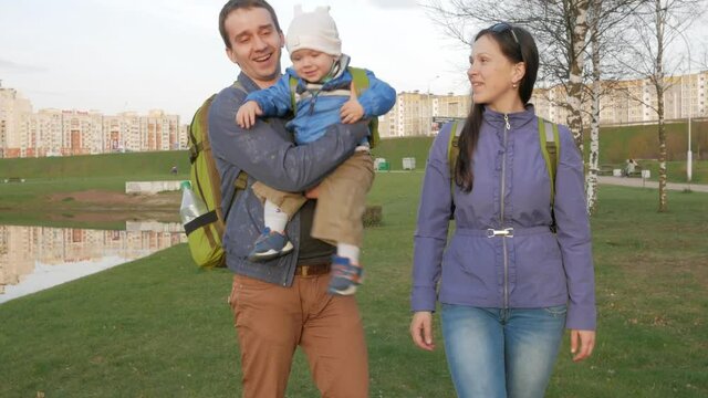 Active family with the baby walks in the park in the spring. Green grass and lake nearby. Green backpacks of different sizes at all